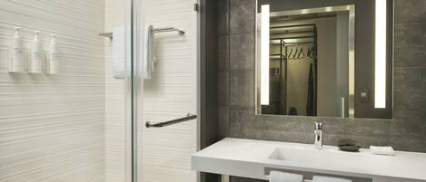 Un baño moderno con mampara de vidrio, lavamanos blanco, baldosas de mármol oscuro y un espejo grande.