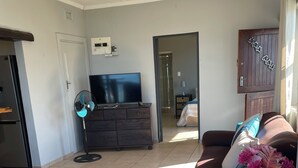 A living room with a sofa, a coffee table, and a TV. There is a door leading to another room and a window with curtains. - Wake up to gorgeous sunrises and sea views upon a wooden balcony. (Amanzimtoti)