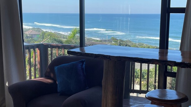 A room with a view of the ocean, a wooden table, a chair, and a small stool. - Wake up to gorgeous sunrises and sea views upon a wooden balcony. (Amanzimtoti)