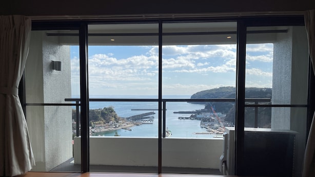 Habitación con un amplio ventanal corredera que da a un balcón con vistas a la costa, incluyendo barcos y un muelle.