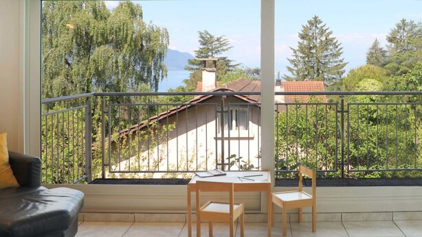 Un balcón con vista a una casa y árboles. Se observa un sofá de cuero negro y una mesa de madera con sillas.