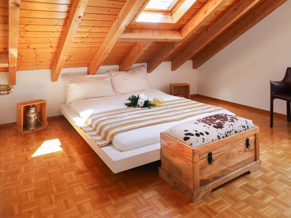 Un dormitorio con techo inclinado, vigas de madera, una cama con manta a rayas, un cofre de madera y una mesita de noche con una vela.
