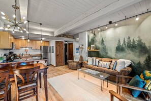 A spacious living area with a dining table, chairs, a sofa, and a coffee table. There is a kitchen area with wooden cabinets and a refrigerator.