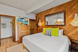 A bedroom with a wooden ceiling, a bed with white bedding, a dresser, a mirror, and wall-mounted lamps.