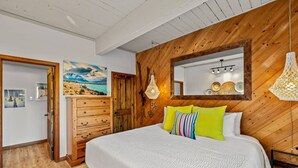 A bedroom with a wooden ceiling, a bed with white bedding, a wooden dresser, a mirror, and a painting on the wall.