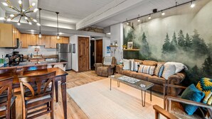 A spacious living area with a large couch, wooden dining table, and chairs. There is a kitchen in the background with wooden cabinets and modern appliances.