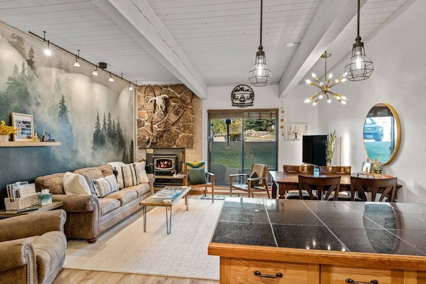 A cozy living room with a fireplace, a stone wall mural, and a dining area with a large window.