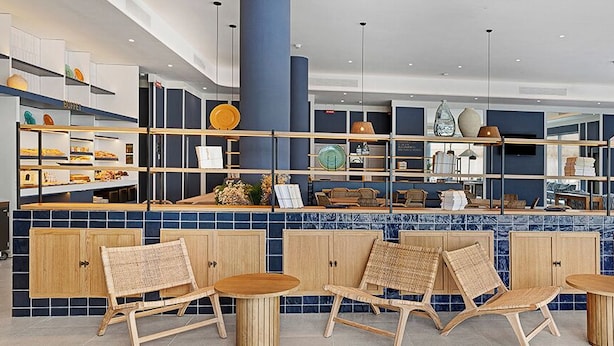Un café moderne aux couleurs bleu et blanc, avec des chaises et des tables en osier, et une variété d’objets décoratifs disposés sur des étagères.