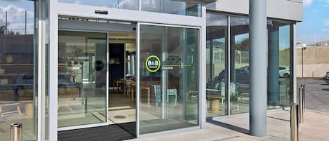 Une entrée d’hôtel moderne avec des portes vitrées, un comptoir de réception bien visible et le logo de la chaîne B&B Hotels.