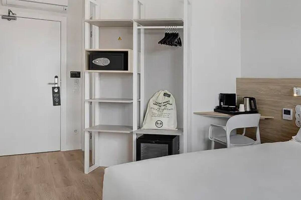 A hotel room with a bed, a desk with a chair, a wardrobe, and a small trash bin.