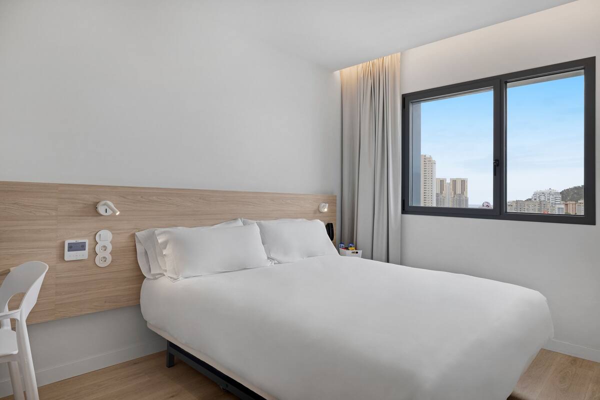 A modern hotel room with a bed, a desk, a chair, a window with a city view, and a control panel on the wall.