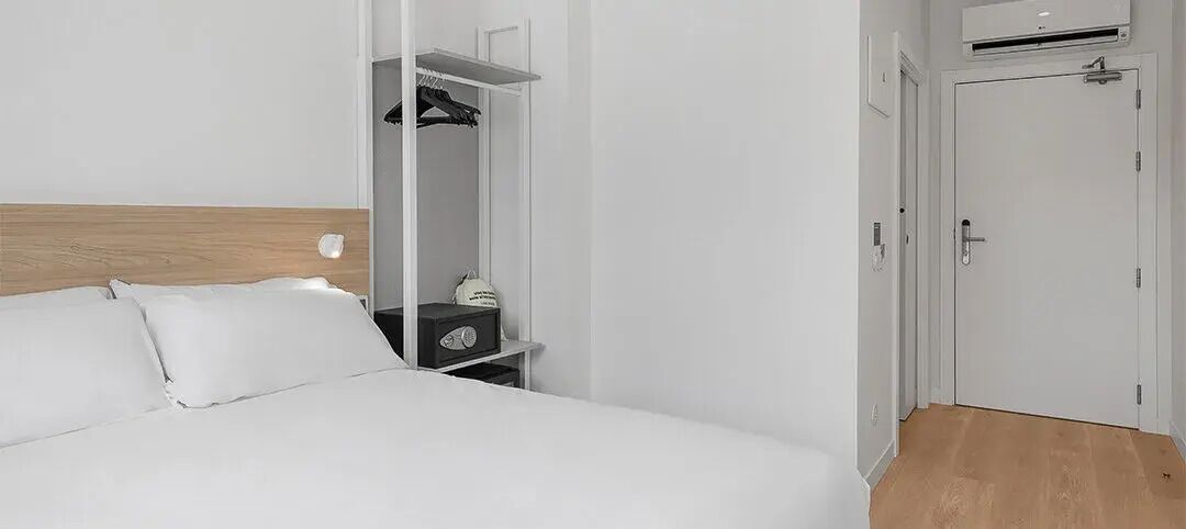 A hotel room with a bed, a wardrobe, a bedside table, and an air conditioning unit.