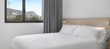 Habitación de hotel con una cama grande, una ventana con vistas a edificios y colinas, y un cabecero de madera.
