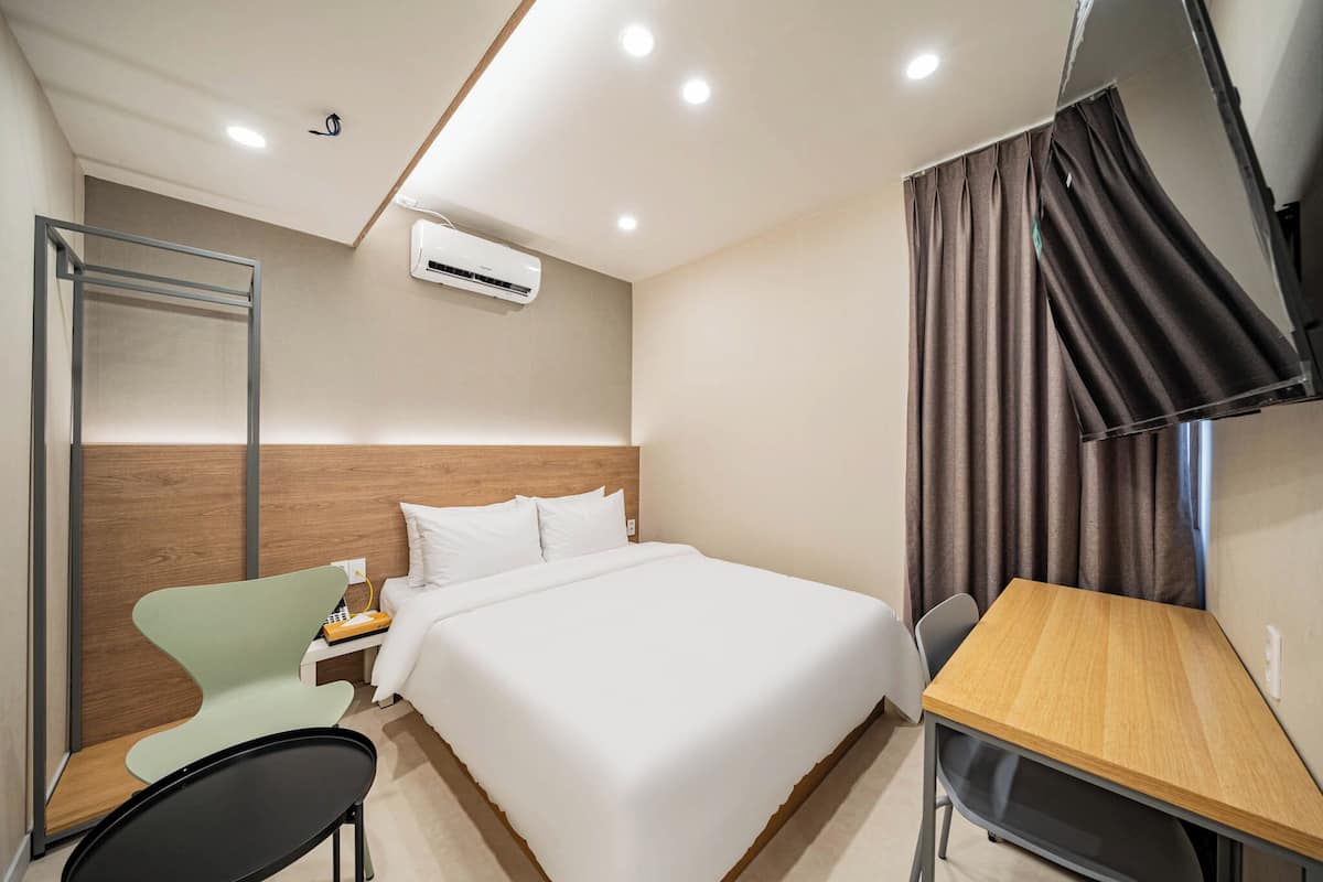 A hotel room with a bed, a desk, a chair, an air conditioning unit, and a curtain.