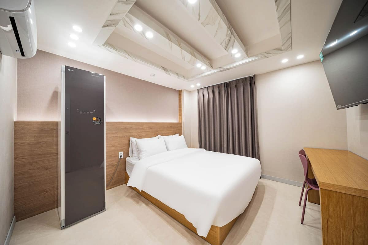 A hotel room with a bed, a desk, a flat-screen TV, and a modern air conditioning unit.