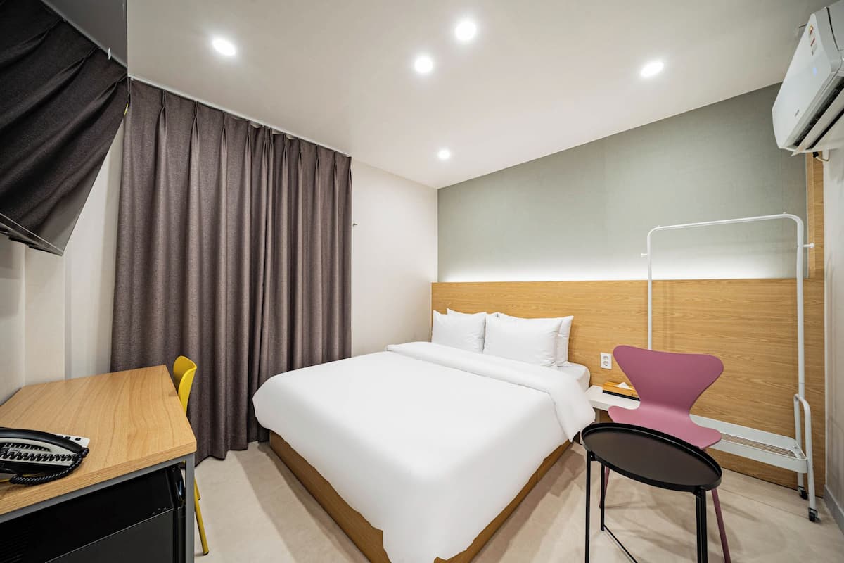 A hotel room with a bed, a desk with a telephone, a chair, a bedside table with a book, and an air conditioning unit.