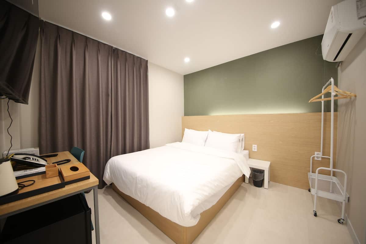 A hotel room with a bed, a desk with a lamp, a chair, a bedside table, a trash bin, and a coat rack.