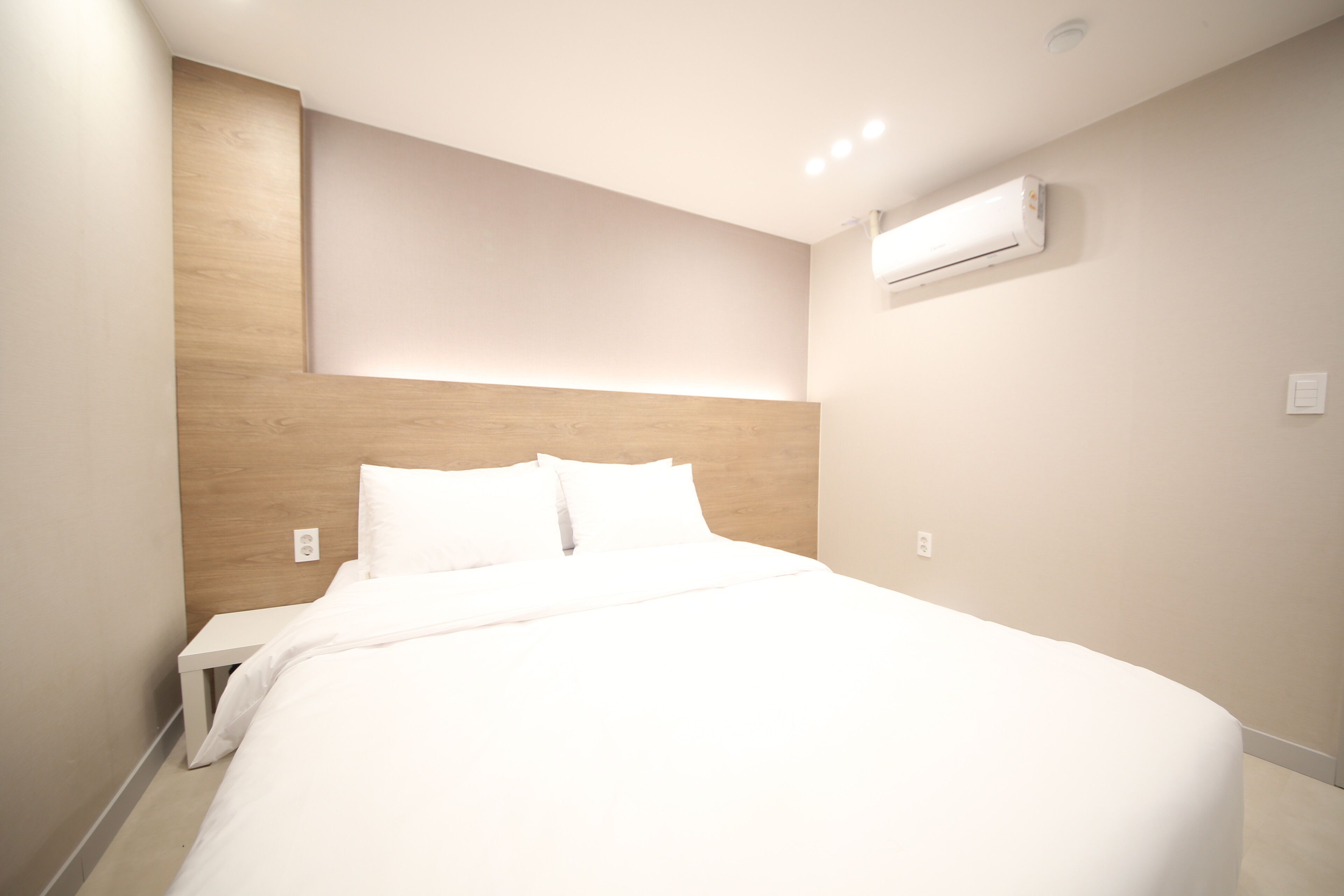 A modern hotel room with a large bed, a wooden headboard, a small bedside table, and an air conditioning unit on the wall.