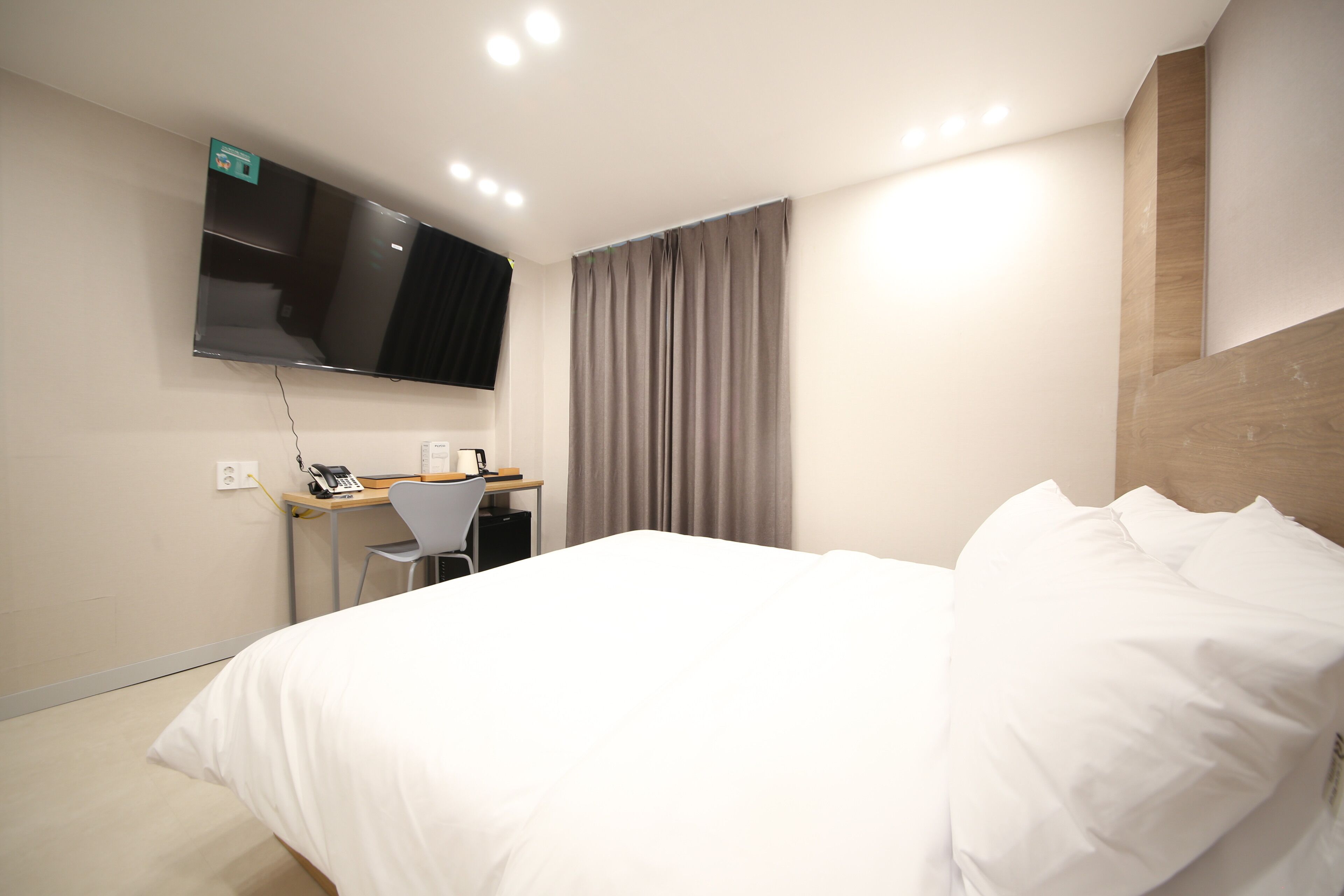 A hotel room with a bed, a desk with a chair, a flat-screen TV mounted on the wall, and a window with curtains.