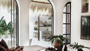 Un salon accueillant avec un canapé, une table ronde en bois sur laquelle est posé un saladier rempli de fruits, et des plantes en pot. Une grande fenêtre offre une vue sur un coin repas et une cuisine en arrière-plan.