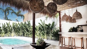 Un espace aménagé au bord de la piscine, avec une table en bois, des suspensions en osier et une végétation tropicale luxuriante.