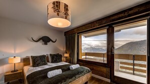 Een slaapkamer met een groot bed, een houten hoofdeinde, een opgehangen dierenhoofd, een nachtkastje, een lamp en uitzicht op bergen door een schuifdeur.