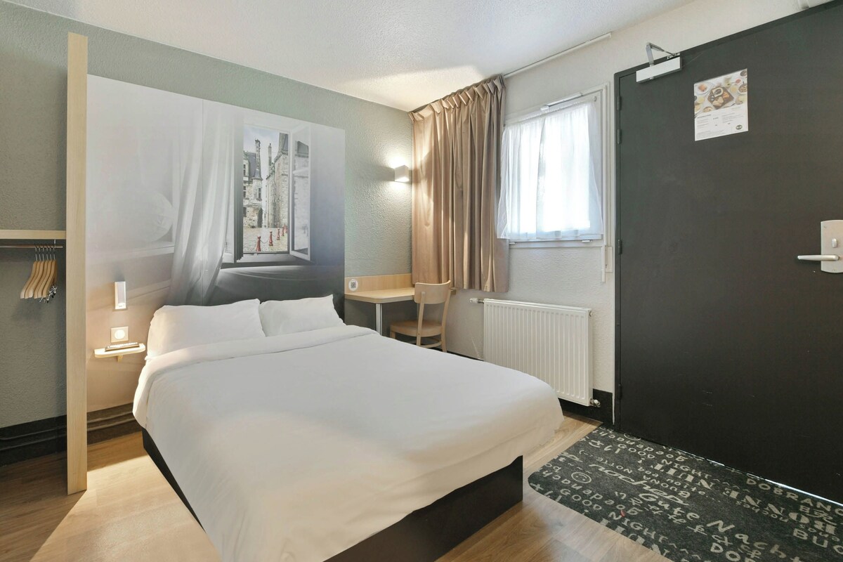 A hotel room with a bed, a desk, a chair, a radiator, a door, and a window with curtains.