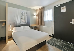 A hotel room with a bed, a desk, a chair, a radiator, a door, and a window with curtains. - B&B Hotel Le Mans Sud (Arnage)