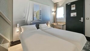 Ein Hotelzimmer mit zwei Betten, einem Schreibtisch, einem Stuhl, einem Fenster mit Vorhängen und einer Tür mit einem Korkbrett.