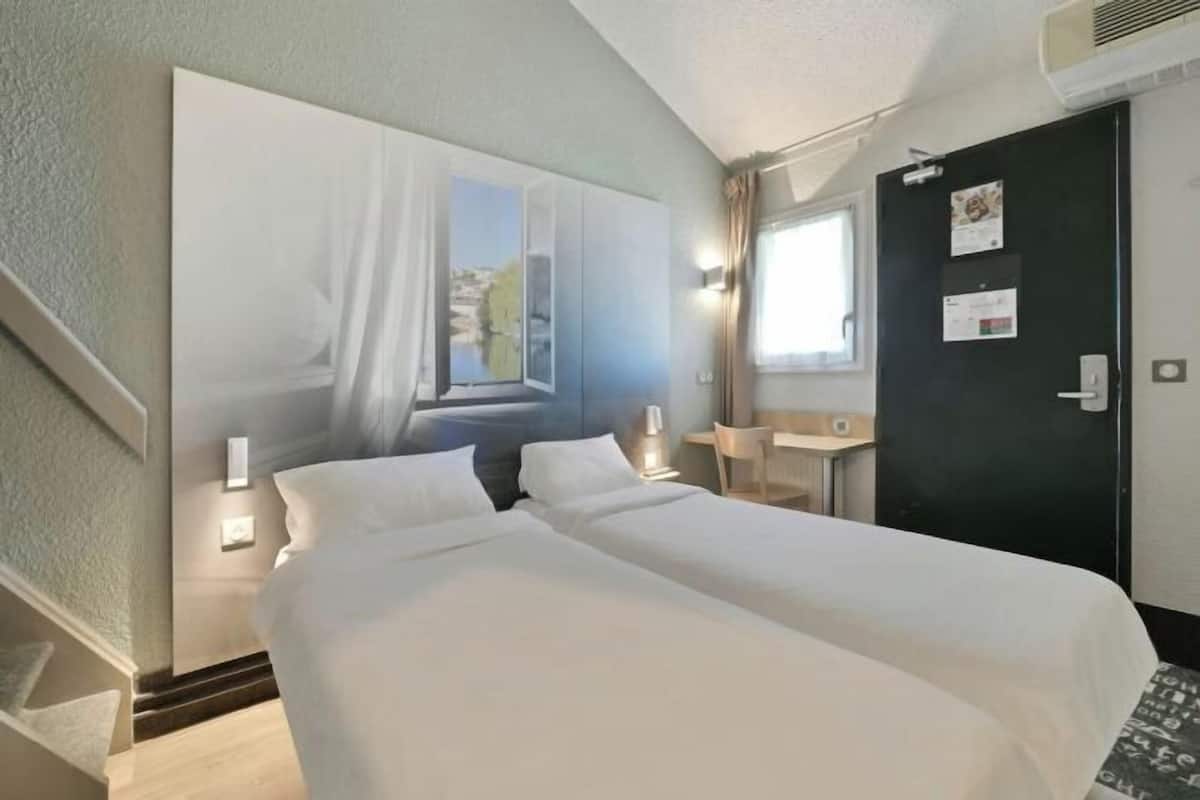 Hotelkamer met twee bedden, een bureau, een stoel, een raam met gordijnen en een deur met prikbord.