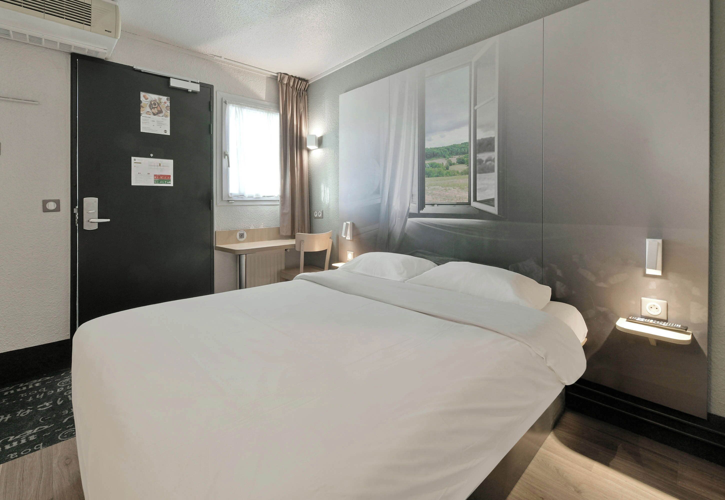 A hotel room with a bed, a desk, a chair, a window with curtains, and a wall-mounted light.