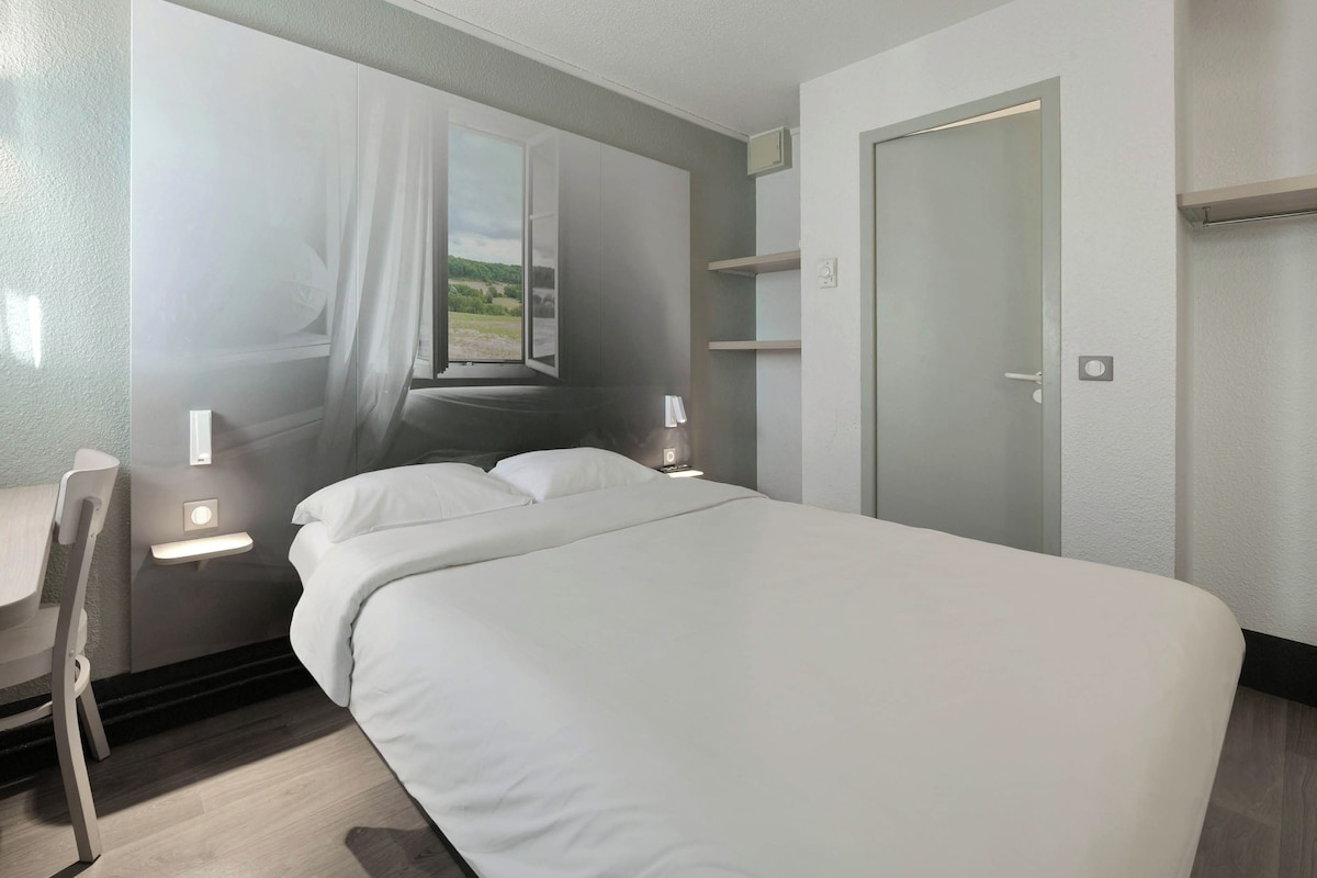 A modern hotel room with a large bed, a desk, a chair, a window with curtains, and a door.