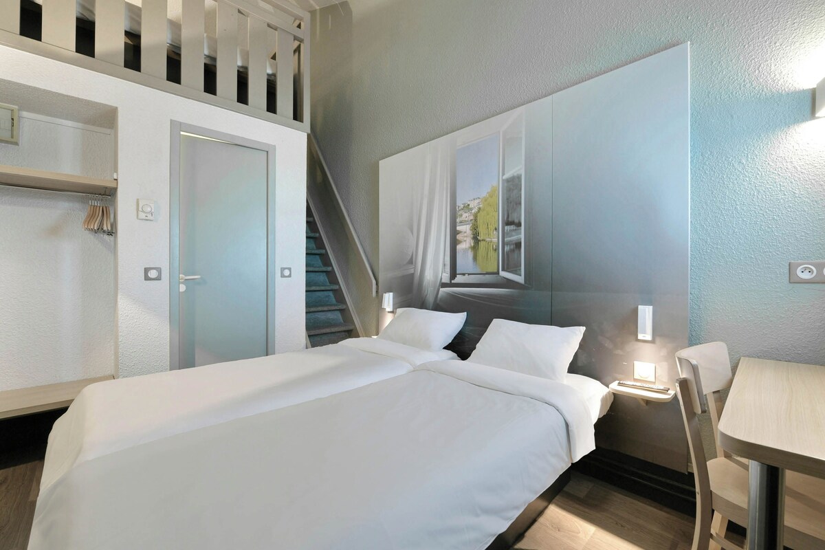 Una camera d'albergo con un letto, una scrivania, una sedia, una scala e un'ampia finestra con vista.