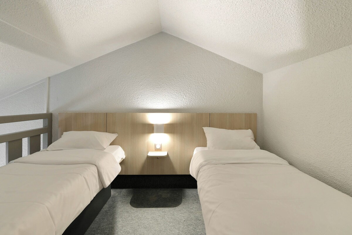A room with two single beds, a wooden headboard, a nightstand with a lamp, and a gray carpet.