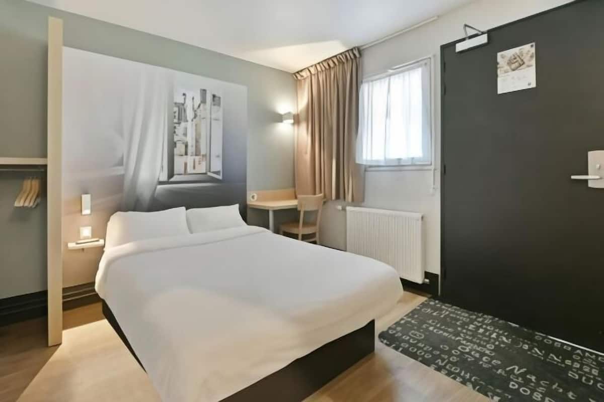 Een hotelkamer met een bed, een tafeltje, een stoel, een radiator, een raam met gordijnen en een muur met een poster.