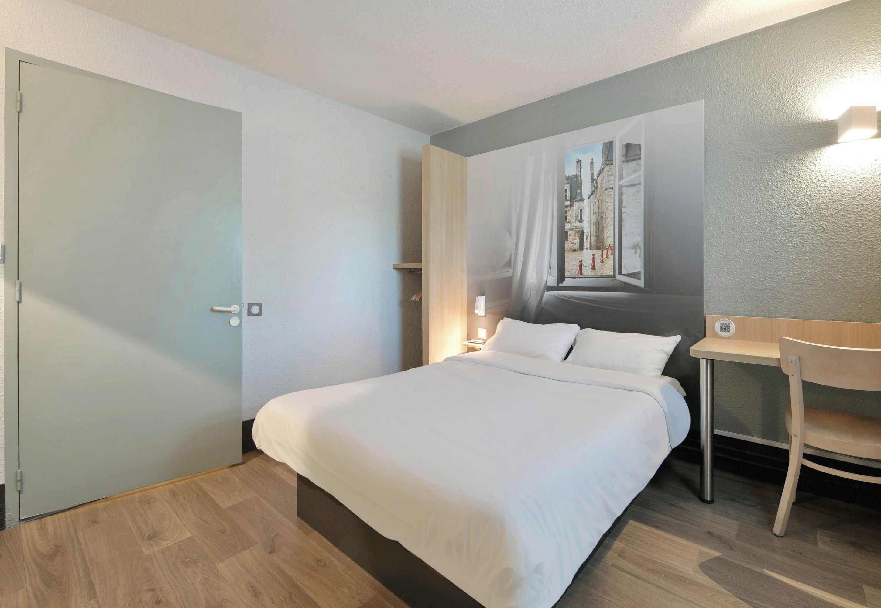 Ein Hotelzimmer mit Bett, Schreibtisch, Stuhl, einem Bild an der Wand und einer Tür.
