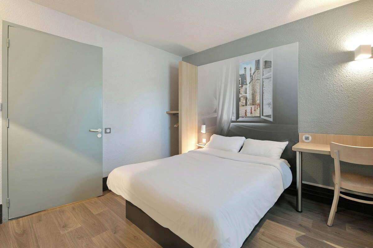 Une chambre d’hôtel avec un lit, un bureau, une chaise, un tableau au mur et une porte.