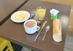 A breakfast table with a plate of scrambled eggs, a cup of hot chocolate, a glass of orange juice, a loaf of bread, a yogurt container, and a packet of Le Gall chocolate. - B&B Hotel Le Mans Sud (Arnage)