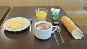 Un plateau de petit-déjeuner avec des œufs brouillés, un verre de jus d’orange, une tasse de chocolat chaud, un pot de yaourt nature, une baguette et des couverts, le tout posé sur une table en bois.