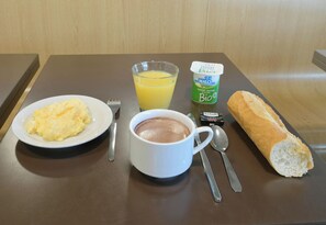 Un plateau de petit-déjeuner avec des œufs brouillés, un verre de jus d’orange, une tasse de chocolat chaud, un pot de yaourt nature, une baguette et des couverts, le tout posé sur une table en bois.
