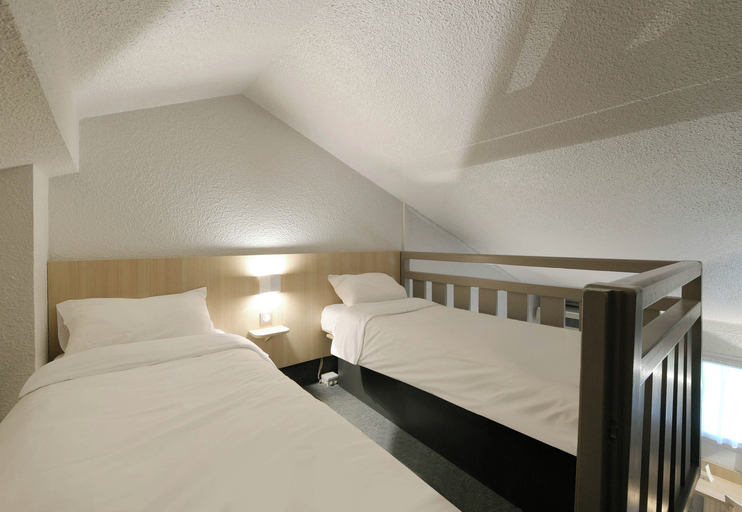 A room with two beds, a nightstand, a wall-mounted lamp, and a sloped ceiling.