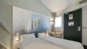 Ein Hotelzimmer mit zwei Betten, einem Schreibtisch, einem Stuhl, einem Fenster mit Aussicht und einer Tür, an der ein Kalender hängt.