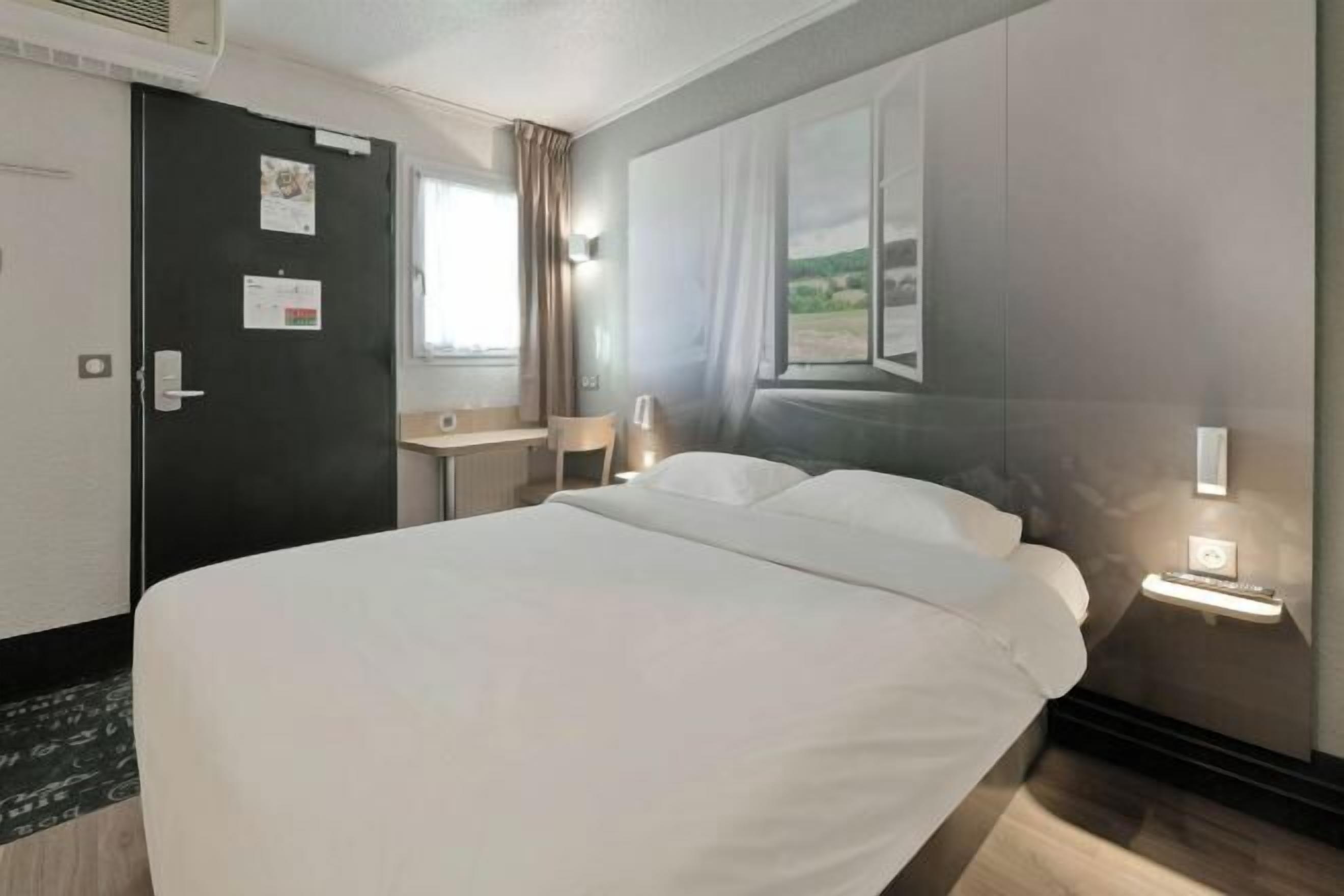 Una habitación de hotel con una cama grande, un escritorio, una silla, un ventanal con cortinas y una mesita de noche fijada a la pared con una lámpara.