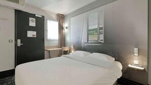 Ein Hotelzimmer mit einem großen Bett, einem Schreibtisch, einem Sessel, einem Fenster mit Vorhängen und einem an der Wand befestigten Nachttisch mit Lampe.