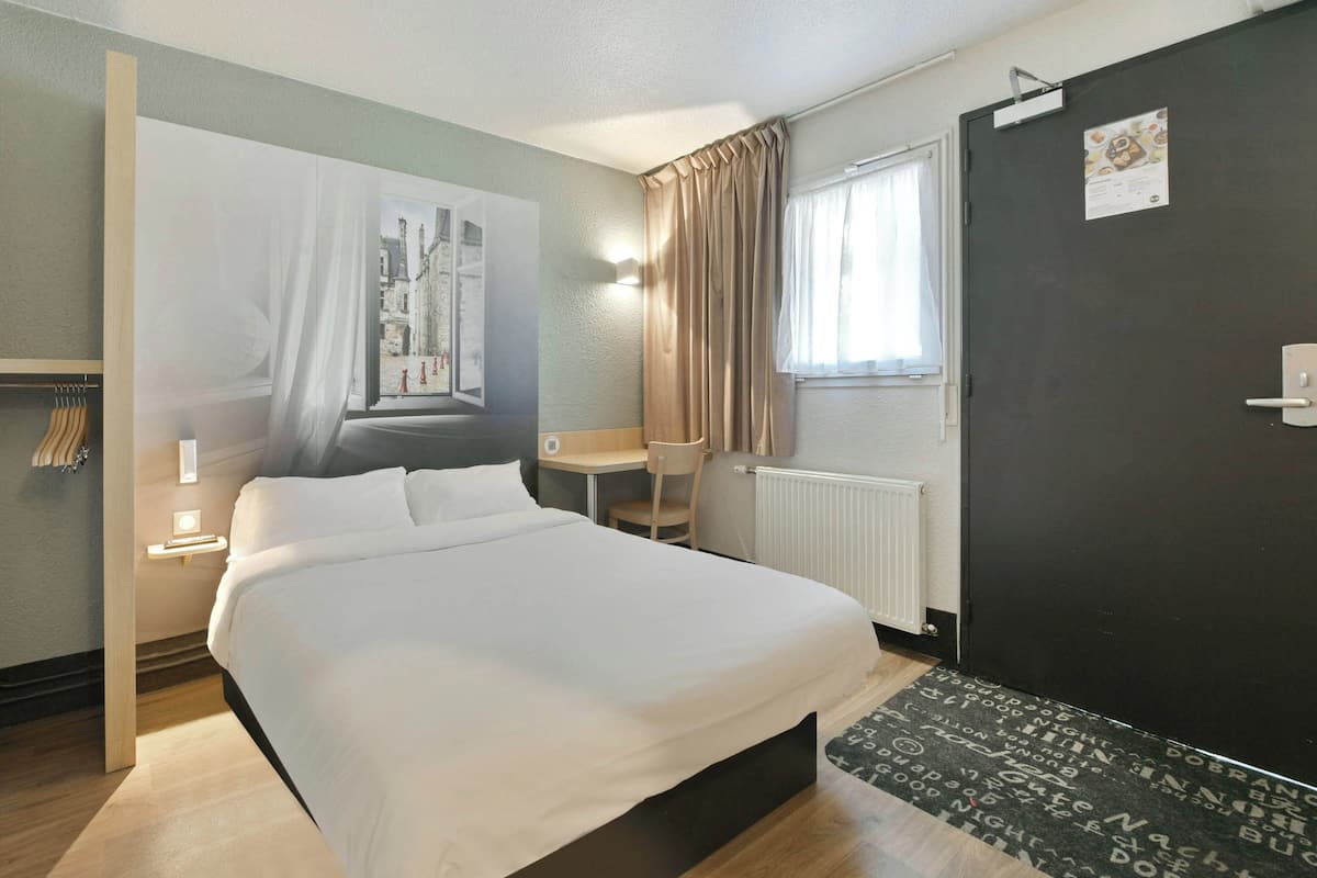 Een hotelkamer met een bed, een bureau, een stoel, een radiator, een deur en een raam met gordijnen.