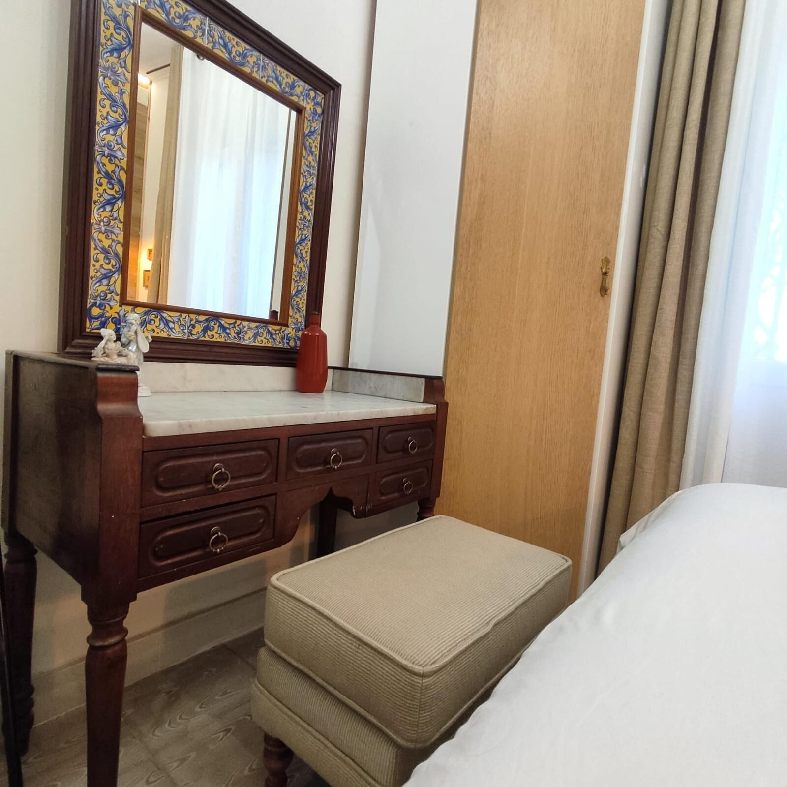 Habitación de hotel con cómoda de madera, espejo con marco decorativo, una pequeña estatua y cama con colcha color beige.