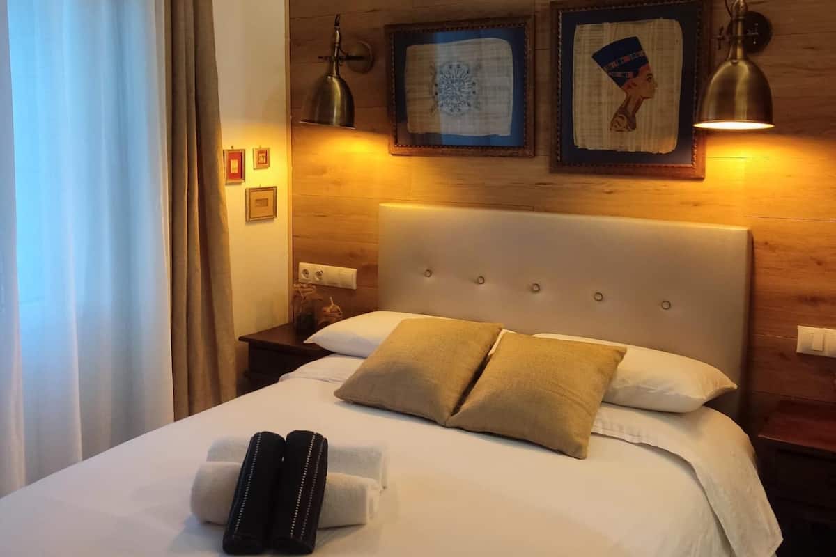 A neatly made bed with two pillows, a nightstand with a lamp, and two framed pictures on the wall.