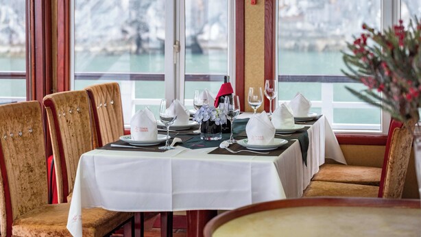Zona pranzo con tavolo imbandito per quattro persone, ampie finestre vista su un corpo d'acqua e sedie imbottite.