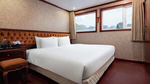 Un letto grande con testiera imbottita, un comodino in legno con un barattolo di vetro e una tazza, una panca e una finestra con tende.
