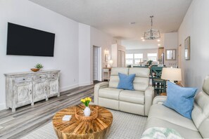 A spacious living room with a large flat-screen TV, a white sofa set, a wooden coffee table, and a dining area in the background. - Beachy, Brand New & Open for Summer! Family Home 5 min. Bike to Beach, 15 min. Drive to Downtown! (St. Augustine)